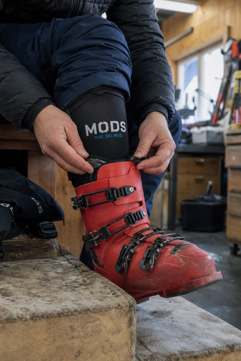 Skier adjusting a red ski boot with a black MODS lower-leg sleeve visible above it.