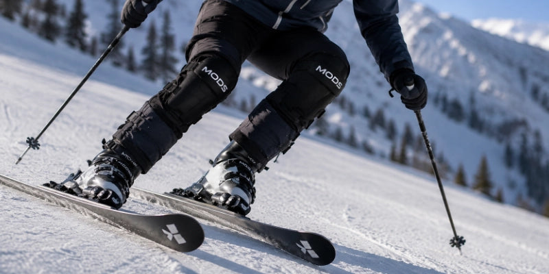 Close-up of a skier leaning into a downhill turn with MODS leg supports fitted over black ski trousers.