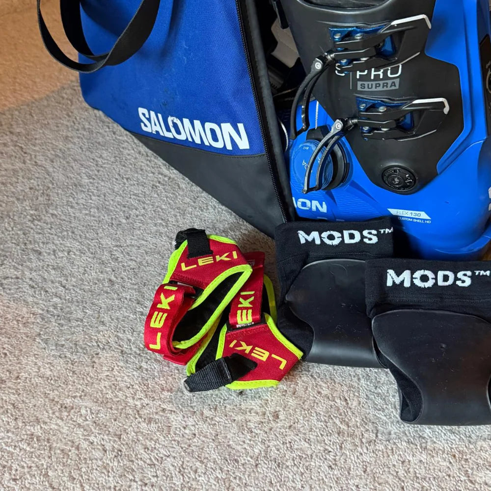 Pair of MODS ski shin protection sleeves positioned with ski boots, poles and equipment, ready to provide targeted cushioning and improved comfort while skiing.