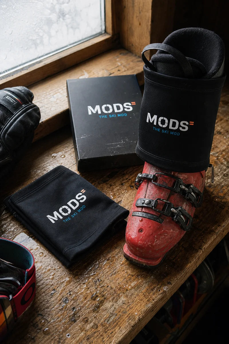 Two black MODS lower-leg sleeves are displayed on a weathered wooden bench beside a frosted window. One sleeve is positioned above a worn red alpine ski boot, while the second is folded next to the MODS product box.