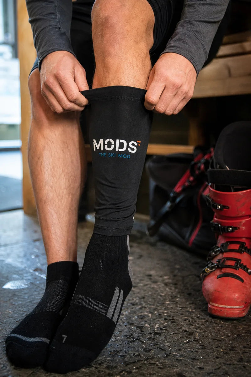 Seated skier pulling a black MODS sleeve over the lower leg beside a red alpine ski boot.