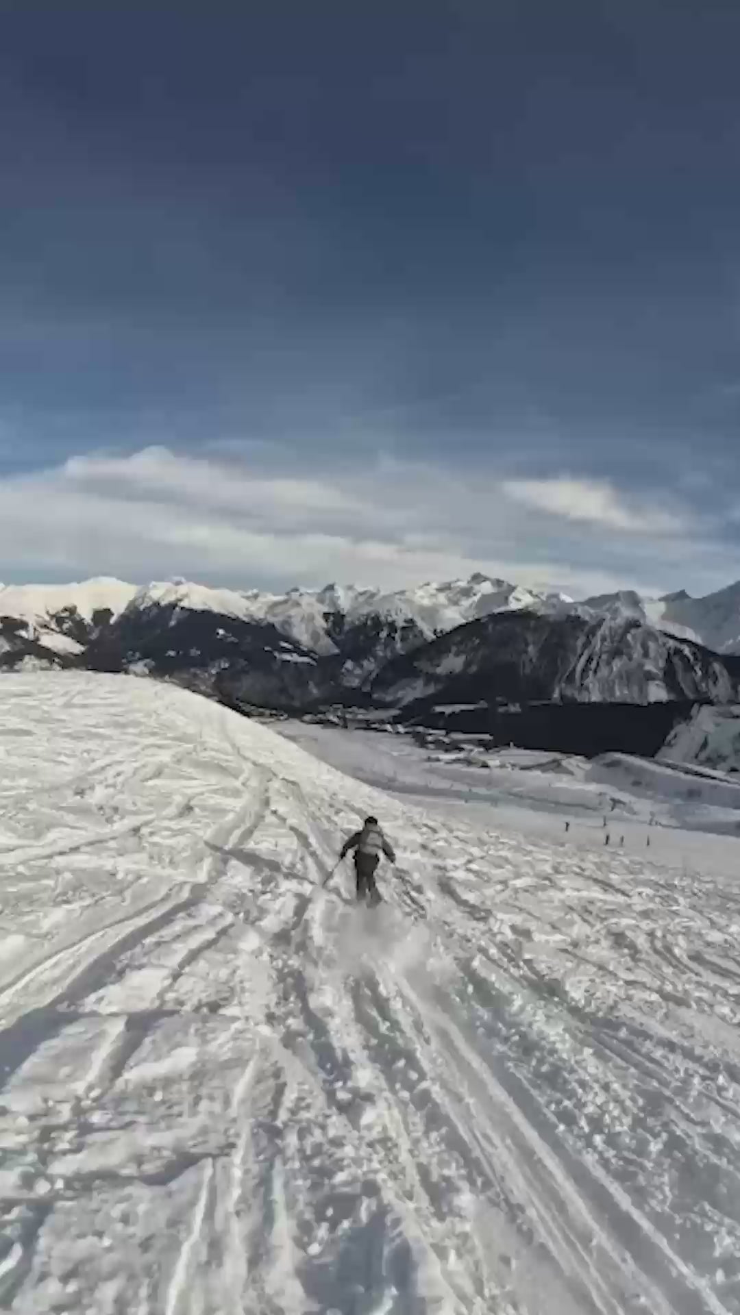 Skier descending a wide snow-covered mountain slope with alpine peaks in the distance.